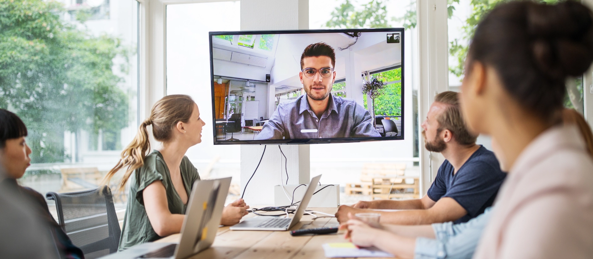 Eine gemischte Gruppe Mitarbeitender der Branche Banking unterhalten sich in einem Remote Meeting mit einem Kollegen, der via Bildschirm zugeschaltet ist