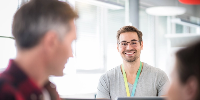Nachwuchsentwickler lächelt inmitten einer Bürosituation über die Bildschirme hinweg in die Kamera