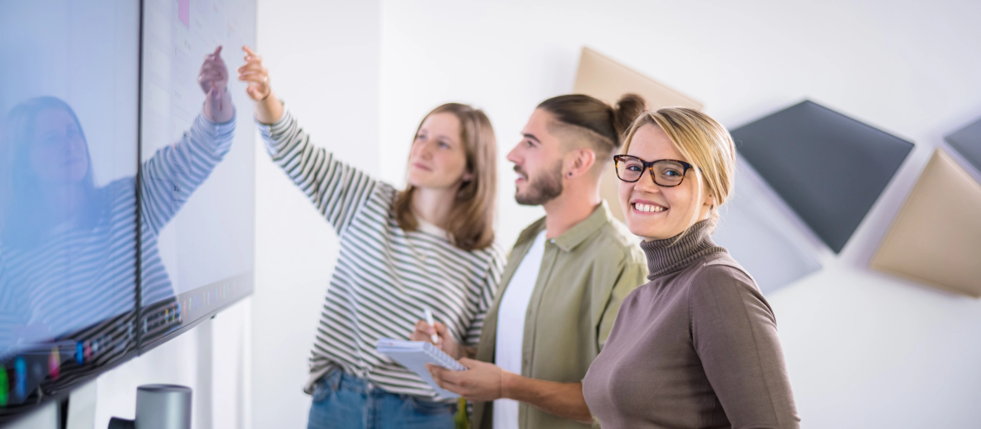 Drei studentische Mitarbeitende stehen vor einem großen Bildschirm und betrachten ein Kanban-Board
