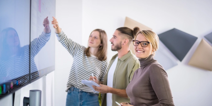 Drei studentische Mitarbeitende stehen vor einem großen Bildschirm und betrachten ein Kanban-Board
