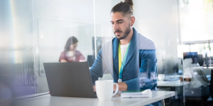 Nachwuchsentwickler sitzt konzentriert in verglastem Büro mit Kaffee an seinem Laptop