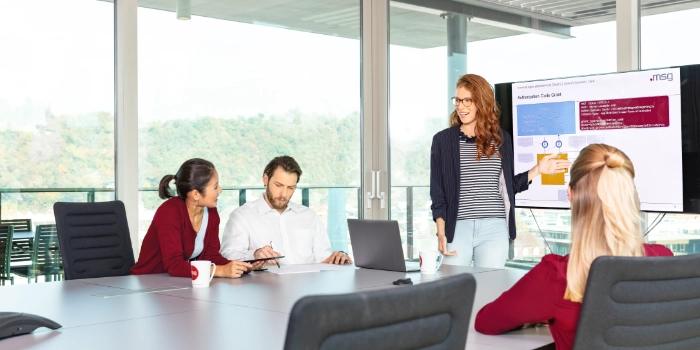 Eine Junior Mitarbeiterin präsentiert in einem Meetingraum etwas ihren Kolleginnen und Kollegen